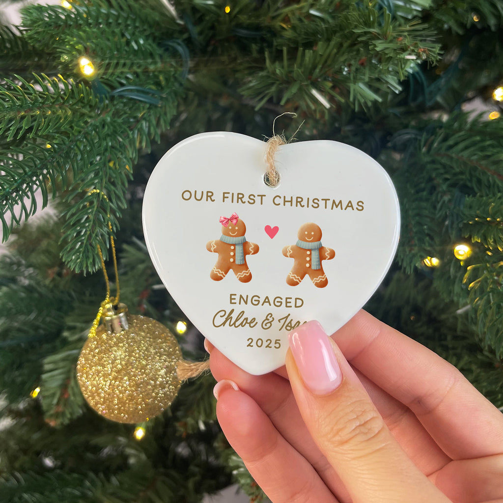 Heart-shaped ornament with 'Our First Christmas Engaged' text and gingerbread men, held in front of a decorated Christmas tree.