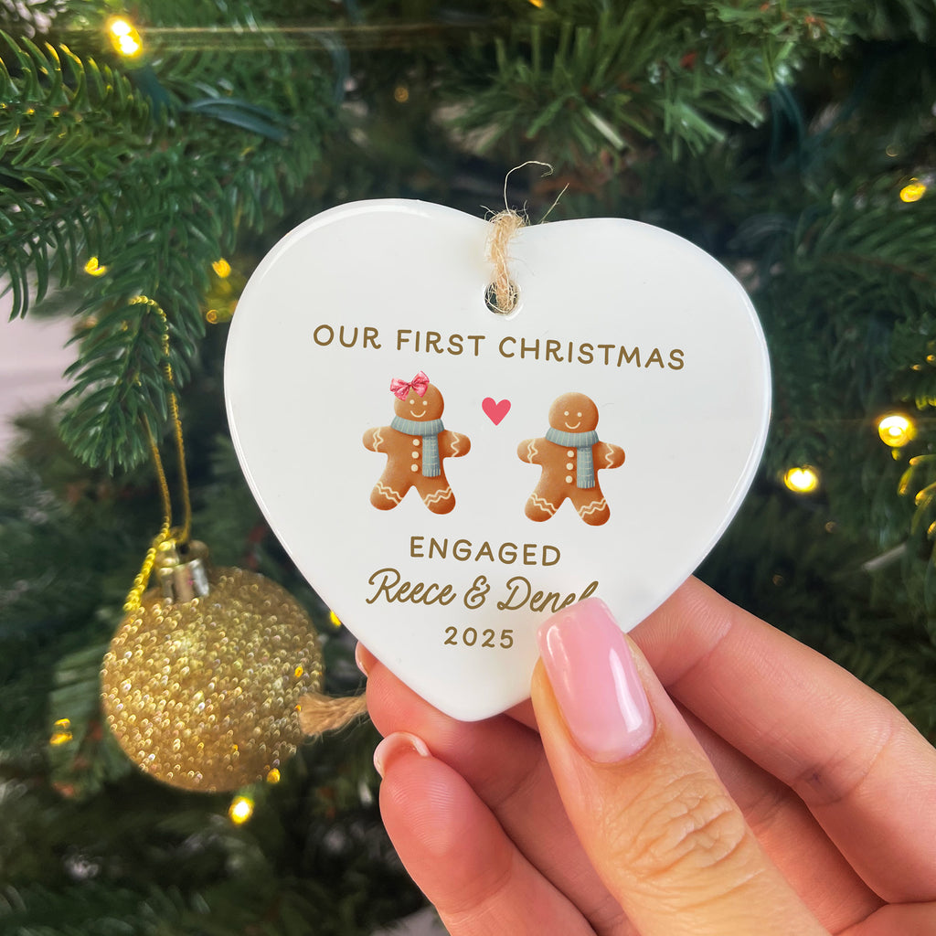 Heart-shaped ornament with 'Our First Christmas Engaged' text and gingerbread men design, held in front of a decorated Christmas tree.